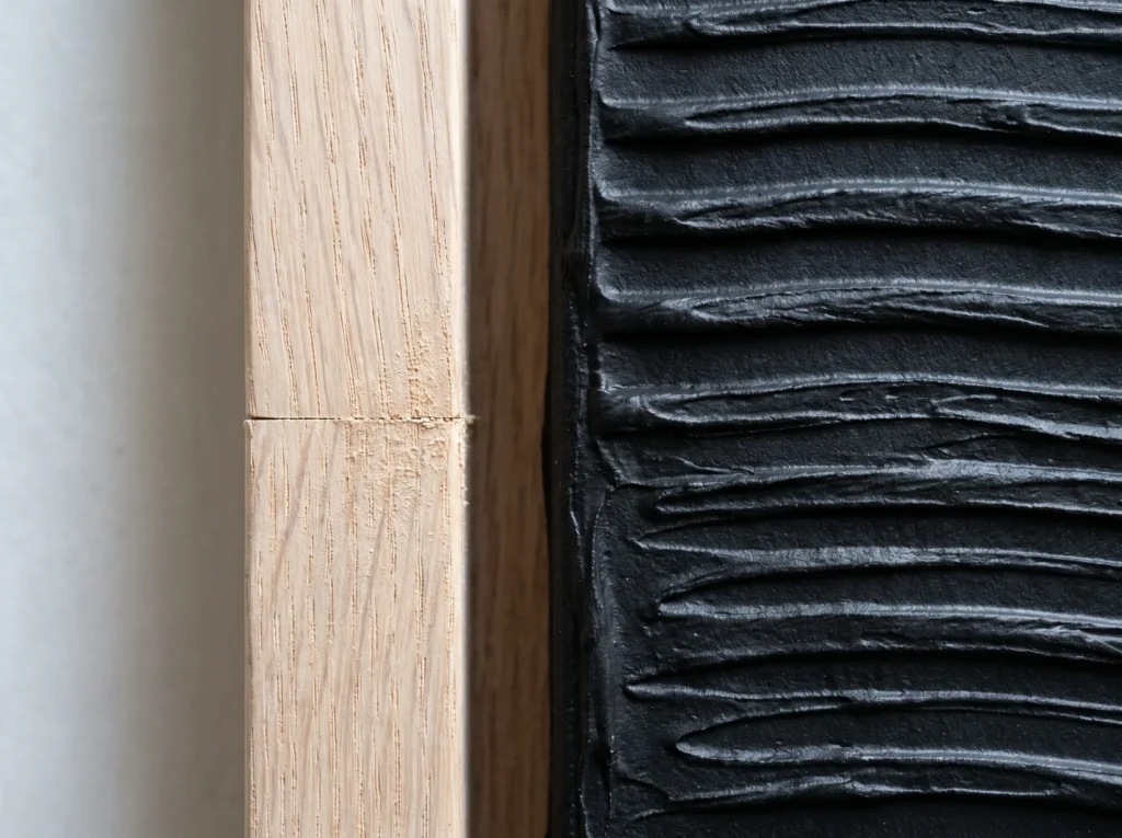 Deep black textured artwork with horizontal ridges is set within a light tan wood frame against an off-white wall.
