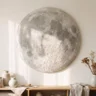 A round plaster moon artwork with realistic crater texture in soft grey-white, hanging on a warm white wall above a low shelf