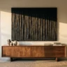 A black plaster wall artwork with deep vertical ridges casting strong shadows in low side light, hanging above a walnut sideboard
