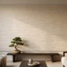A japandi textured wall art panel with calm horizontal ridges in pale oatmeal plaster, hanging above a low dark wood bench with a small bonsai