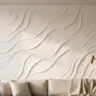 A neutral textured wall artwork in warm white with soft impasto waves, styled above a linen sofa with cushions, a throw and a brass floor lamp beside it