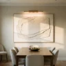 A gallery-grade sculptural textured canvas with deep layered relief in cream and stone, lit by a picture light, hanging in a refined dining room