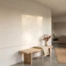 A large minimalist plaster wall mural with soft sweeping trowel ridges in warm white covering a section of a hallway wall, simple wooden bench below