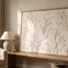 A luxe textured artwork on a canvas board with soft sculpted botanical relief in ivory, framed in thin oak, on a styled shelf