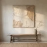 A large wabi sabi plaster artwork with rough, earthy, softly cracked off-white and clay texture hanging on a limewashed wall above a weathered wooden bench with a single dried branch in a stoneware vase