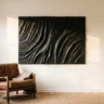 A dramatic 3D black plaster wall artwork with deep ridged texture catching light, hanging on a warm white wall above a leather lounge chair
