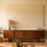 A mid-century modern plaster wall artwork with smooth, flowing parallel wave grooves in warm cream, hanging above a vintage walnut credenza styled with a ceramic lamp and a stack of design books