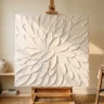 A textured canvas artwork made with spatula strokes forming crisp overlapping petal-like ridges in warm white, on an easel in a bright studio corner