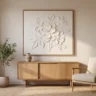 A white sculpted plaster flower relief on canvas in a thin oak frame, hanging above a simple sideboard with a ceramic vase