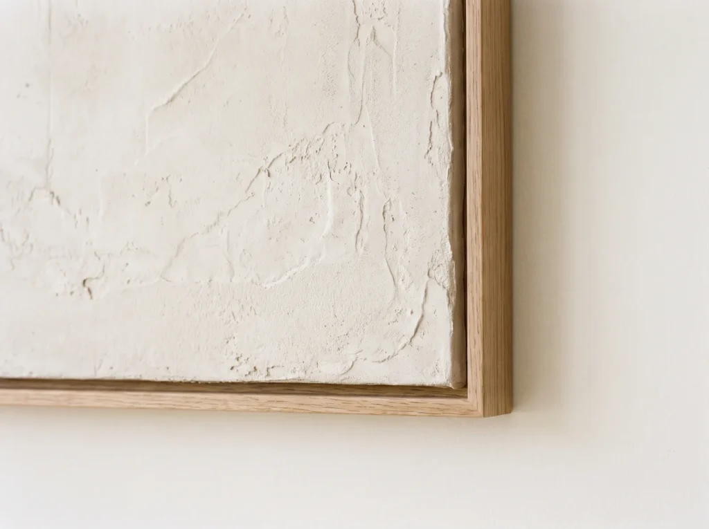 Corner of a neutral textured plaster canvas in a thin oak float frame, showing the shadow gap that makes it look like it floats.