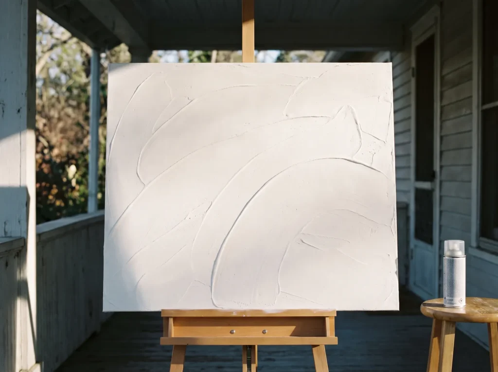 Minimalist white plaster canvas on an easel next to a spray can of clear matte polyurethane on a porch.